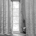 Two grey lined curtains, one with a metallic woven sheen and one with a raised leaf pattern, hanging either side of a tall window with a garden view