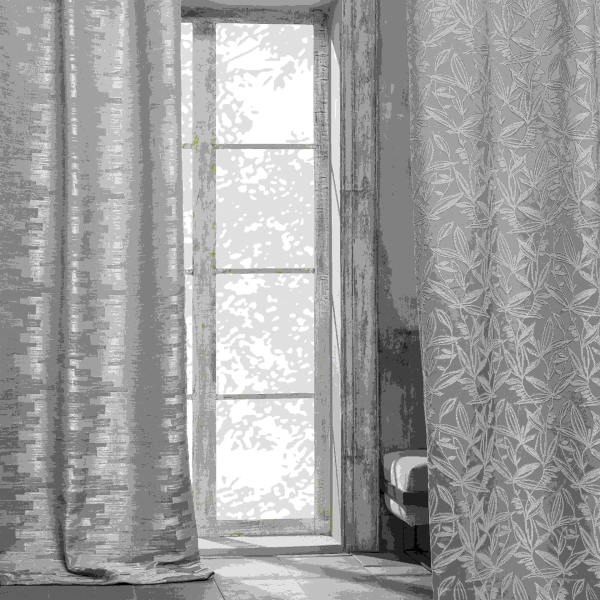 Two grey lined curtains, one with a metallic woven sheen and one with a raised leaf pattern, hanging either side of a tall window with a garden view