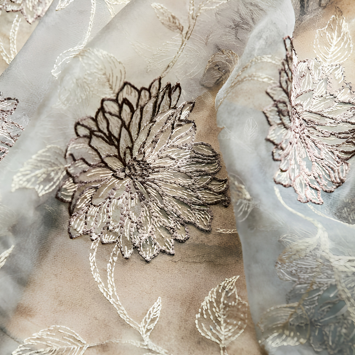 Close-up of embroidered sheer curtain fabric with a brown and cream chrysanthemum motif and leaf outlines in fine thread on grey-blue organza