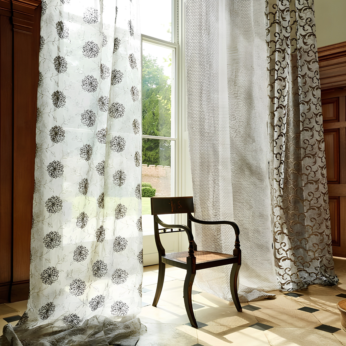 White sheer curtain embroidered with black dahlia flowers beside a tall window with a garden view, a dark wooden chair and patterned curtains alongside