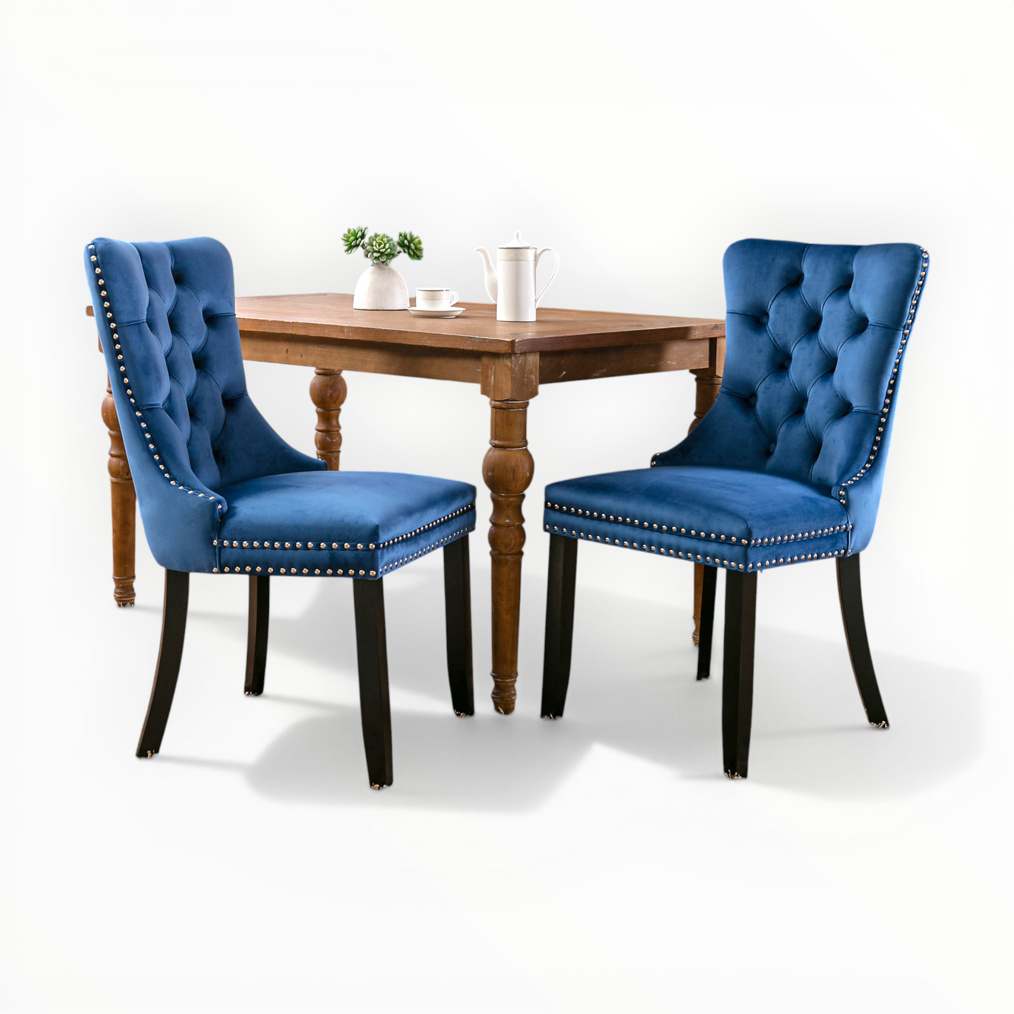 Two blue tufted chairs with nailhead trim in front of a wooden table on a white background