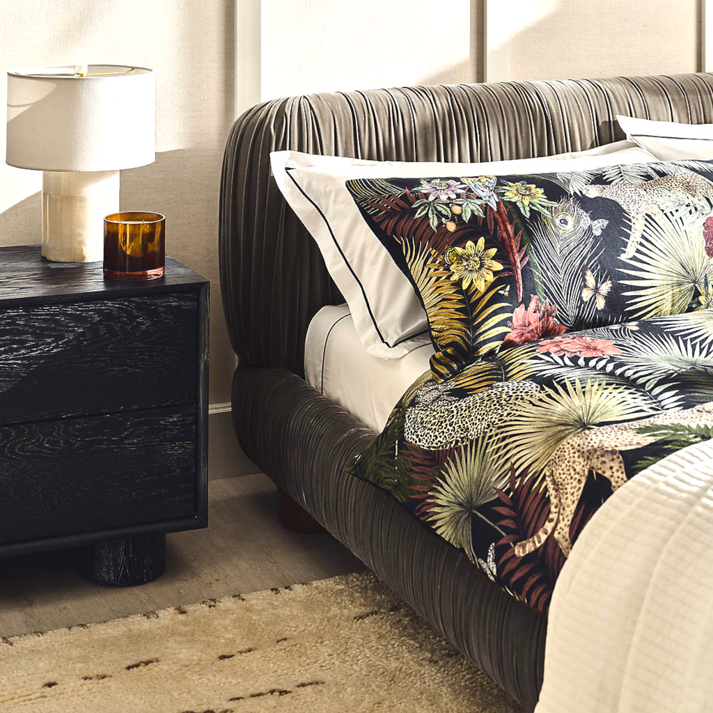 Decorative bed with floral and animal print pillows next to a dark nightstand with a lamp.