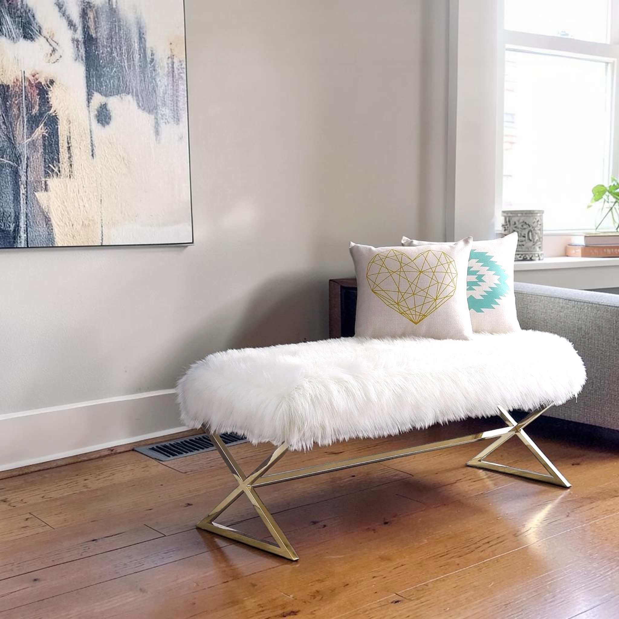 White fur bench with gold legs in a room with a painting on the wall and a window.