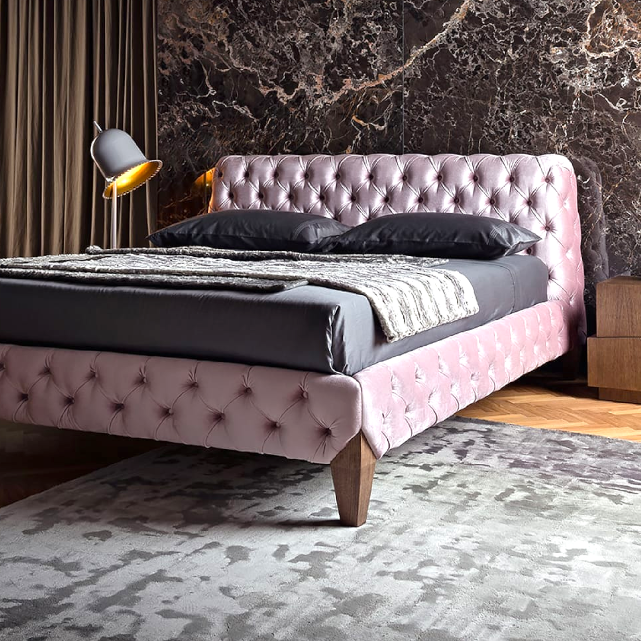 Pink tufted bed with dark bedding in a room with marble wall and lamp.