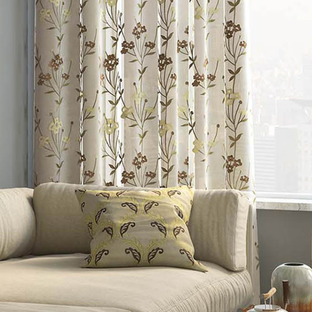 Sheer cream curtain with embroidered brown and yellow floral vines, softly filtering daylight beside a beige sofa with a paisley cushion