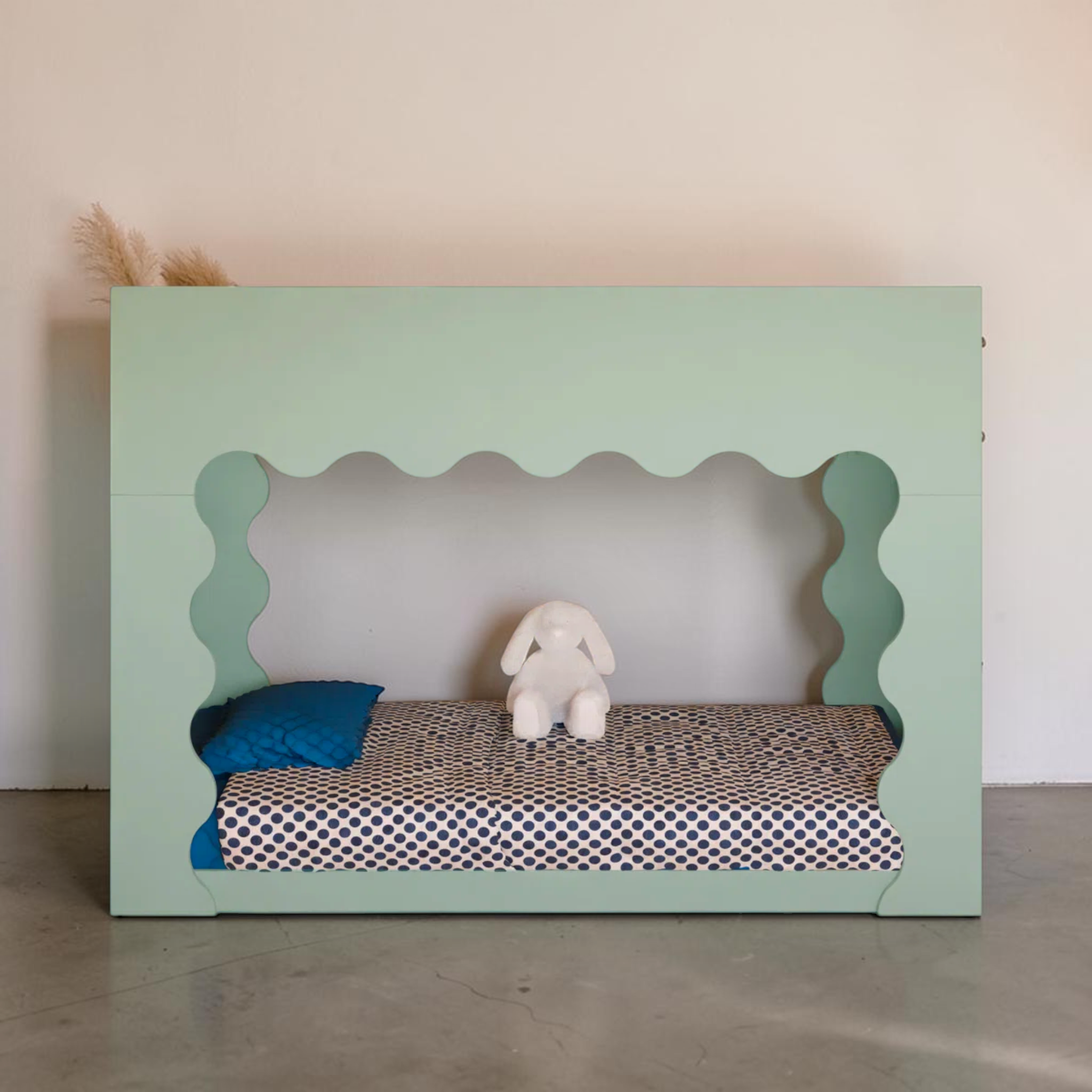 Pet bed with a green frame, blue pillow, and white plush toy on a neutral background