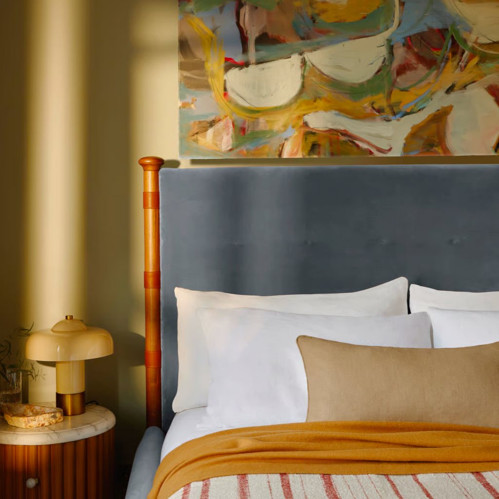 Bedroom with a bed featuring white pillows and a yellow blanket, a nightstand with a lamp, and a colorful abstract painting on the wall.